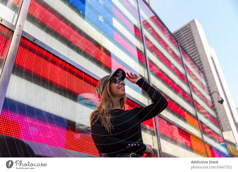 Stylish woman in front of digital American flag in Times Square style sunglasses smile screen Manhattan New York vibrant city street urban fashion cap black