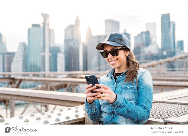 Woman using smartphone with Manhattan backdrop woman sunglasses cap smile skyline cityscape New York outdoor urban bridge fashion leisure casual denim sunset
