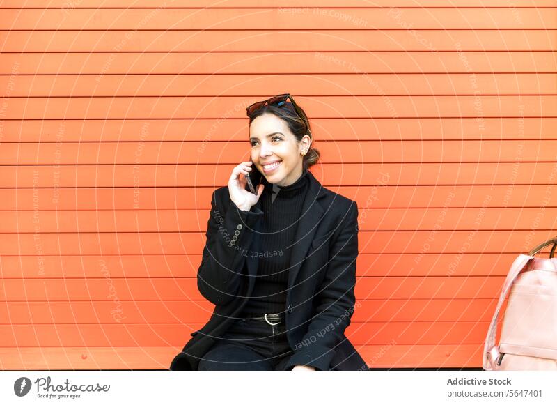 Young woman sitting on orange wall and talking on smartphone phone call speak smile happy street conversation cheerful female young mobile dress device