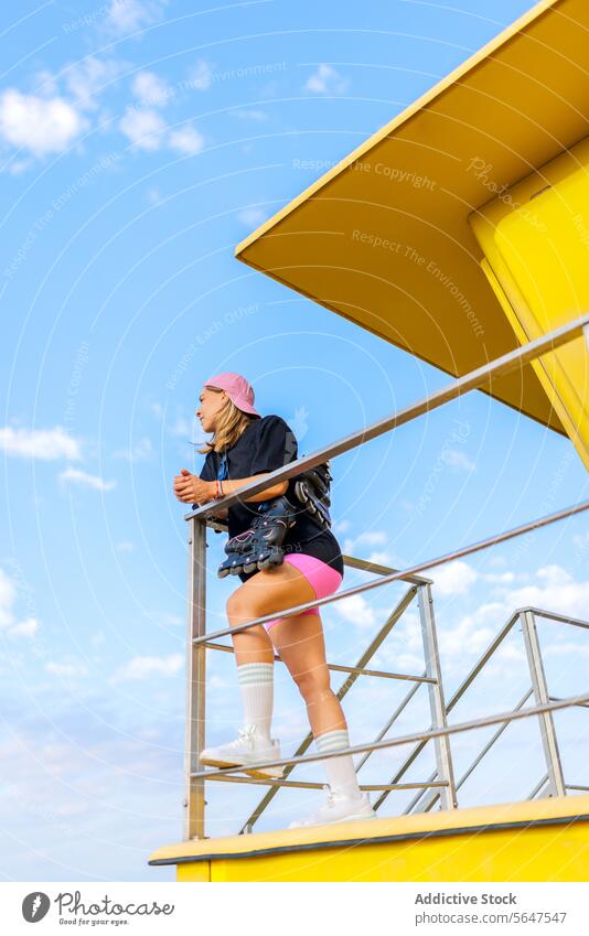 Full length female skater against blue sky woman lifeguard hut beach railing leaning low angle inline roller full length lifestyle weekend leisure hobby