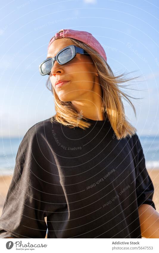 Pretty female at seaside during sunny day woman stylish sunglasses cap beach blond beautiful looking back shoulder pretty fashionable sunlight blur wind tshirt