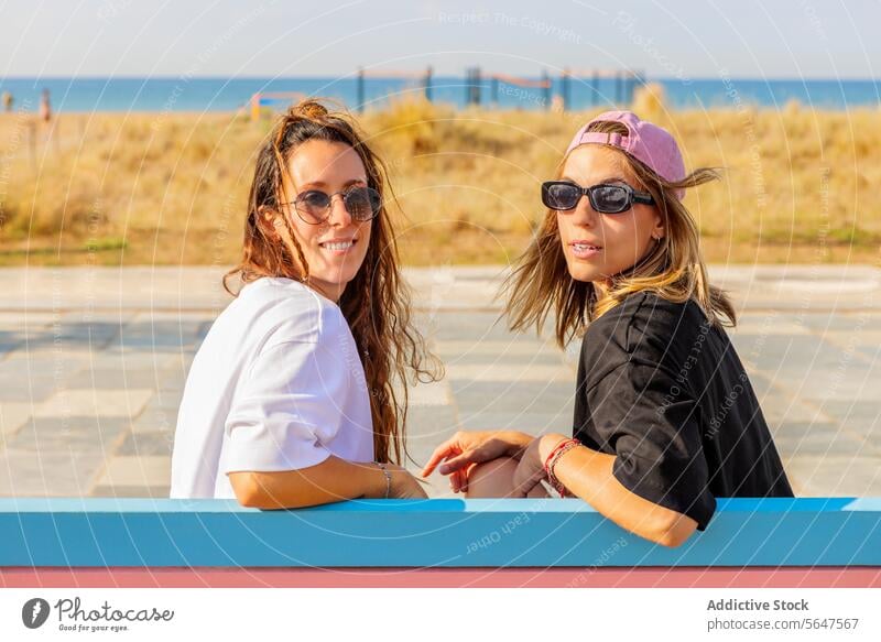 Beautiful women wearing sunglasses during summer portrait friend bench sunny beautiful looking back together lifestyle cap smiling blur sunlight
