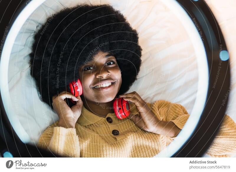 Smiling black woman listening to music with headphones earphones using bed rest home weekend chill female african american afro gadget ring lamp relax device