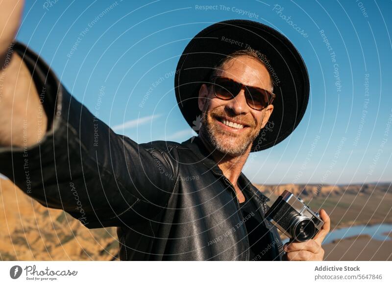 Smiling Traveler Taking Selfie with Camera on Adventure adventurer selfie photographer camera smiling travel exploration desert scenic happy cheerful man hat
