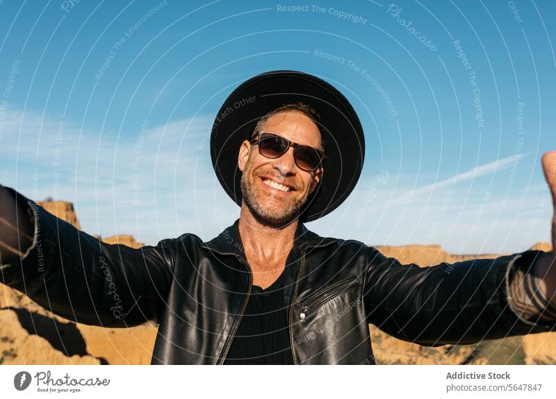 Joyful man taking a selfie with outstretched arms enjoying the outdoors smile happy hat sunglasses black jacket smiling leather blue sky sunshine daytime nature