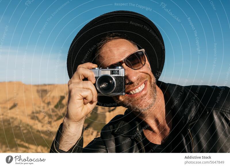 Smiling photographer capturing the moment outdoors with a film camera man smiling vintage hat scenic backdrop joyful taking leather jacket sunglasses happiness
