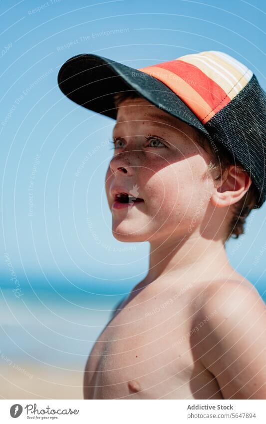 Thoughtful young boy on the beach with baseball cap child contemplative thoughtful sand ocean blue sky swimwear summer sea coastline leisure vacation holiday