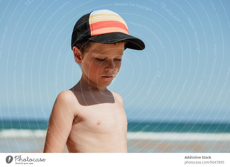 Young boy in thought at the beach child contemplative cap sand shirtless coast young thoughtful summer seascape ocean horizon outdoors daytime youth casual