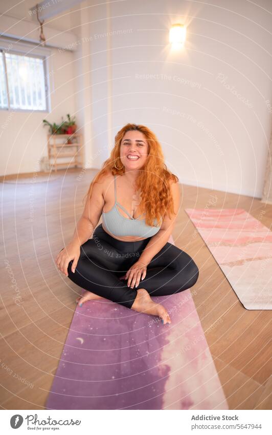 Cheerful curvy woman sitting on mat with crossed legs cheerful activewear smile legs crossed positive lady barefoot leggings top sportswear happy training yoga