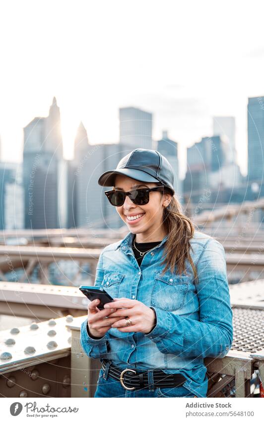 Woman using smartphone with Manhattan backdrop woman sunglasses cap smile skyline cityscape New York outdoor urban bridge fashion leisure casual denim sunset