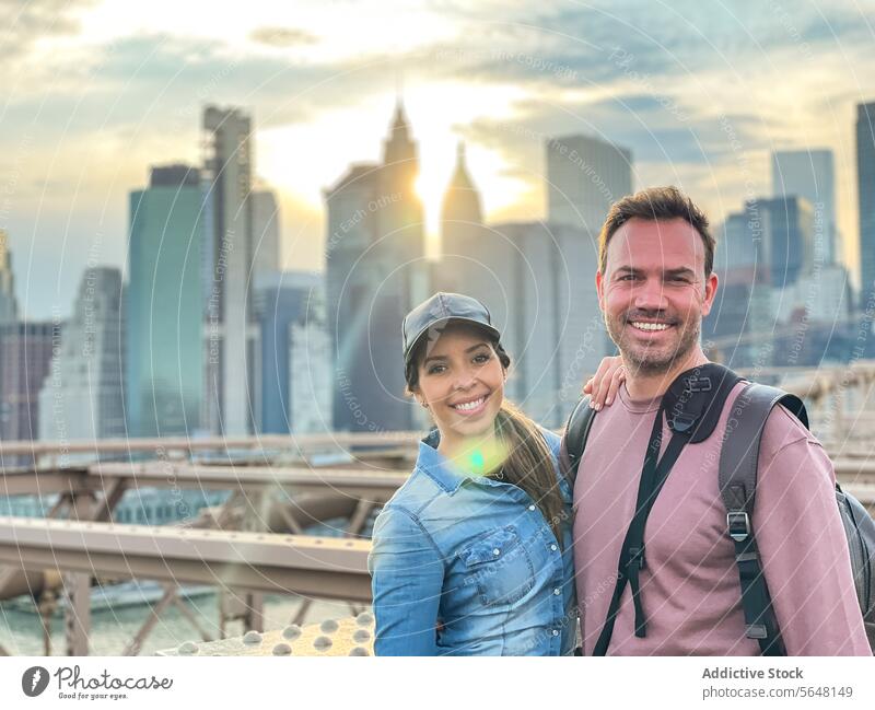 Man and woman bonding with Manhattan backdrop on Brooklyn Bridge skyline sunset New York backpack cap smile couple cityscape skyscrapers travel tourism