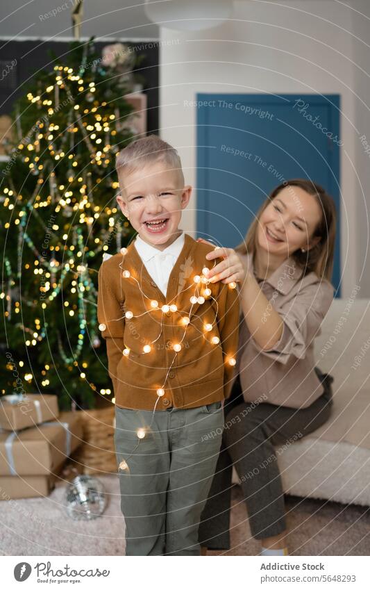 Joyful kid with his mother playing with Christmas lights against Christmas tree at home boy laughter joy woman holiday atmosphere lively affectionate festive