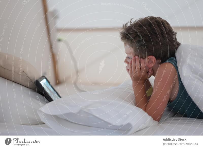 Little boy watching video on tablet while lying in bedroom rest kid internet child blanket relax online using comfort duvet chill digital surfing social media