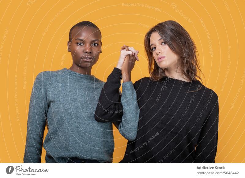 Diverse women in casuals holding hands against yellow background confident friend multiracial young trendy unity together casual attire posing staring sweater