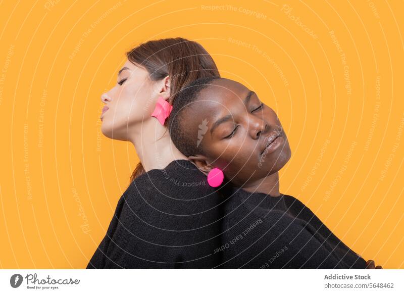 Side view of diverse young female friends leaning on each other with eyes closed while wearing trendy pink earrings against yellow background women together