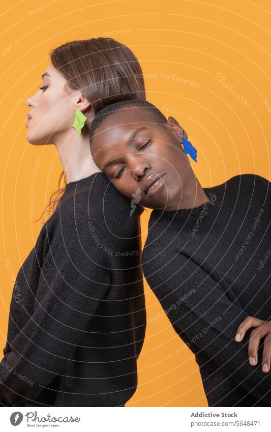 Side view of diverse young female friends leaning on each other with eyes closed while wearing trendy colorful earrings against yellow background women together