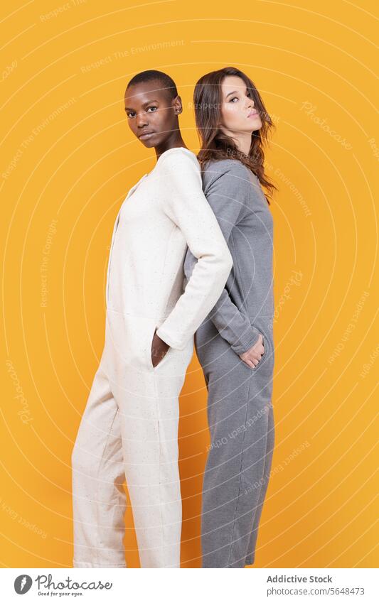 Multiracial women posing with hands in pockets looking at camera confident back to back friend multiracial young trendy together casual attire slim jumpsuit