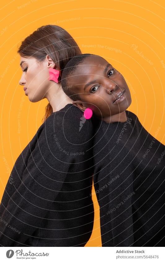 Side view of diverse young female friends leaning on each other looking at camera while wearing trendy pink earrings against yellow background women together