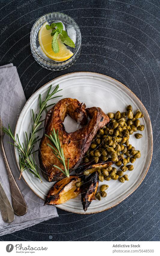 BBQ salmon steak seasoned with spices accompanied by fermented capers, charred lemon, a sprig of rosemary, with a refreshing lemon water on the side grilled