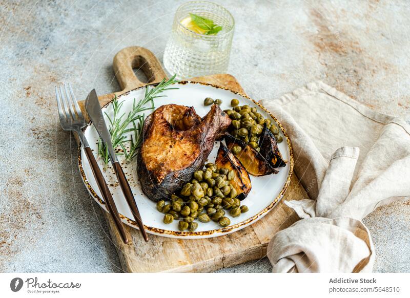 A healthy grilled salmon steak seasoned with spices next to roasted lemons and capers on a wooden board with a linen napkin and lemon water Healthy lunch