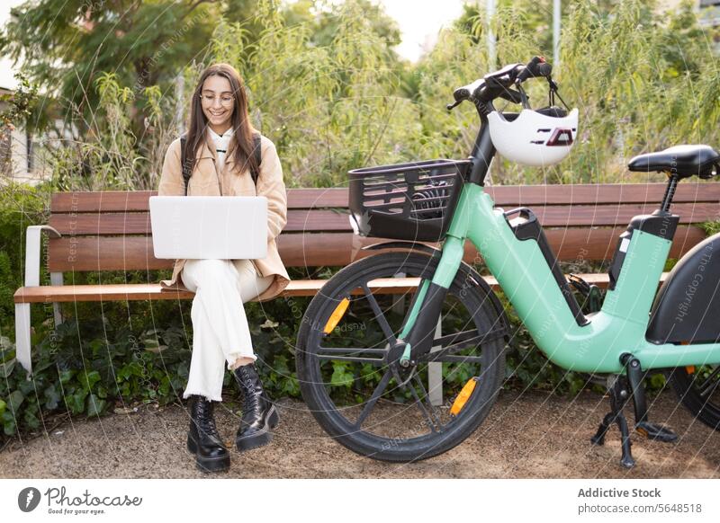 Young woman working on laptop by her electric bike in park bench sunny outdoor typing seated casual wear leisure technology mobile office work-life balance