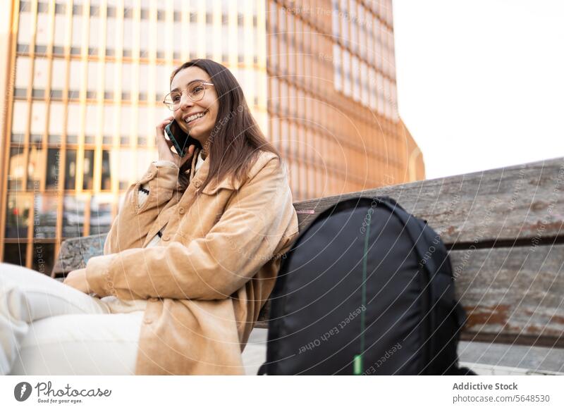 Brunette woman with glasses sitting on a bench and talking on the phone smile cellphone outdoor young adult female city path sunlit cheerful transportation