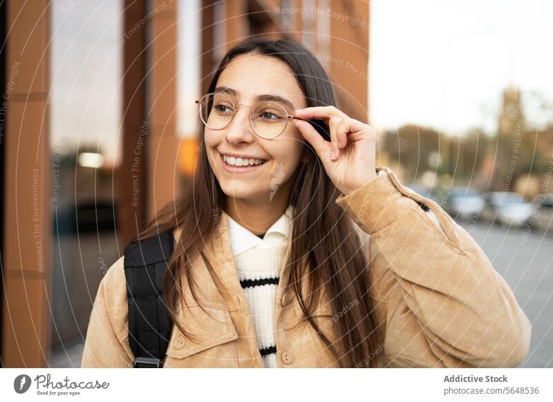Smiling young woman adjusting her glasses on city street smile sidewalk style confidence cheerful urban casual coat beige female fashion happy lifestyle outdoor