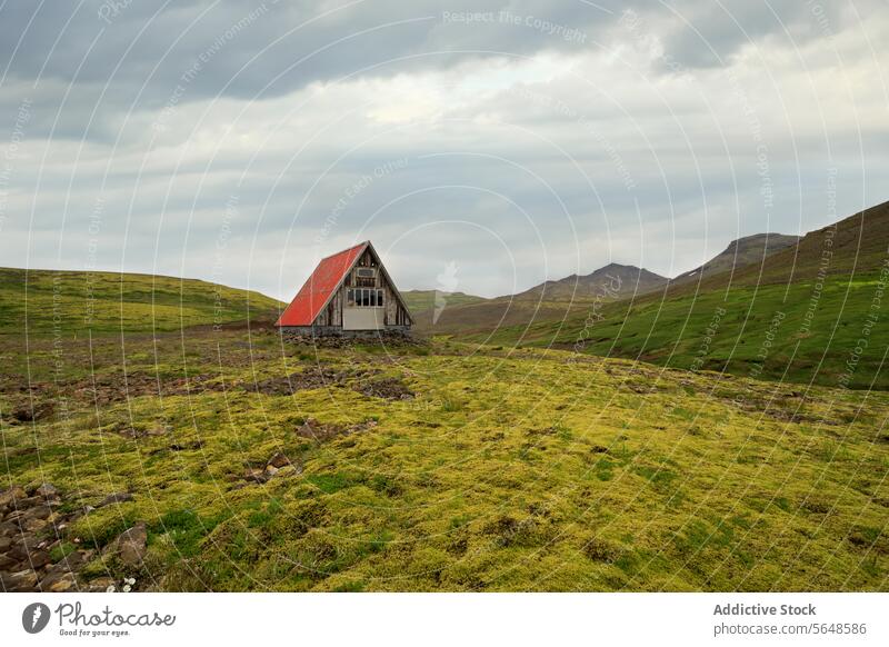 From below of solitary cottage with a red roof stands amidst a vast Icelandic landscape with green hills under a cloudy sky nature outdoor scenic remote
