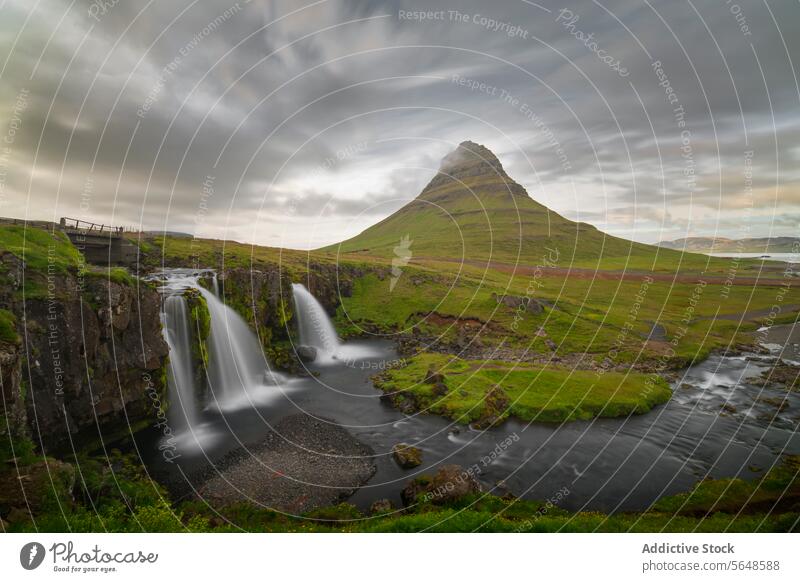 Lush green landscape with a distinctive mountain peak towering over dual waterfalls under a dramatic sky in Iceland nature outdoor scenic lush picturesque