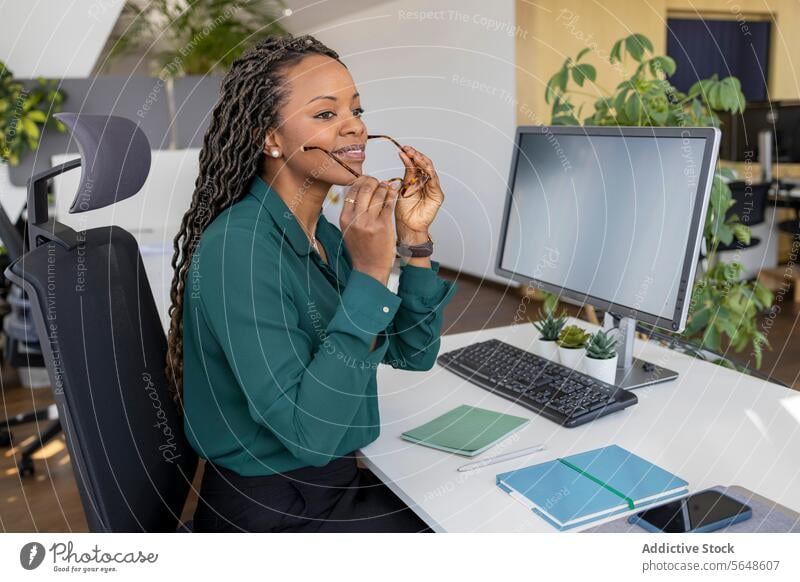 Businesswoman putting on eyeglasses at computer desk in office Eyeglasses Put On Computer Desk Office Workplace Afro Monitor Copy Space Entrepreneur Occupation