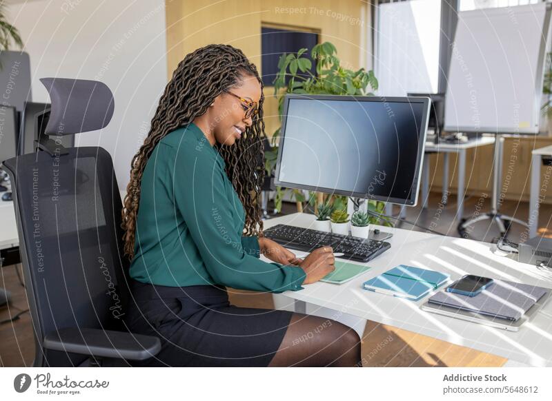 Side view of young happy African American businesswoman writing on notepad at computer desk in office Businesswoman Write Notepad Desk Computer Keyboard