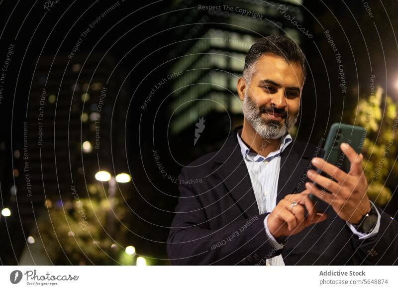 Happy Indian businessman in elegant suit using smartphone on city street cellphone smile beard standing happy texting streetlight illuminate building male night