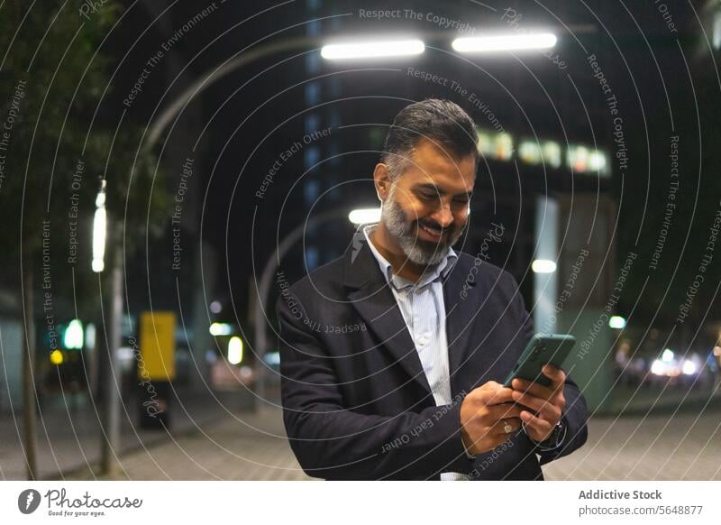 Happy Indian businessman in elegant suit using smartphone on city street cellphone smile beard standing happy texting streetlight illuminate building male night