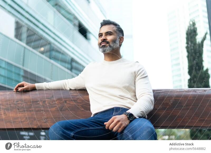 Indian businessman sitting on wooden bench and looking away casual outfit beard indian alone lean on park entrepreneur male urban from below low angle confident