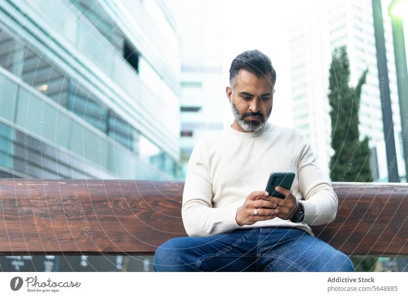 Focused Indian businessman messaging via cellphone while sitting on on street bench message focused smartphone jeans sweater using city concentrate surfing