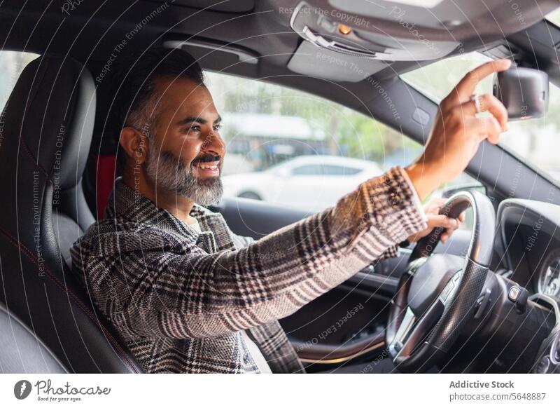Smiling ethnic male entrepreneur sitting in car while looking at rear-view mirror man driver businessman steering wheel automobile road trip vehicle seat
