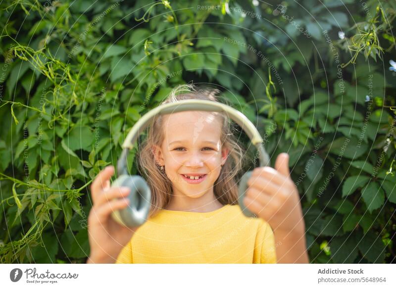 Smiling girl with headphones outdoors against greenery smile thumbs up cheerful young dress yellow holding garden leaf nature summer happiness child kid music