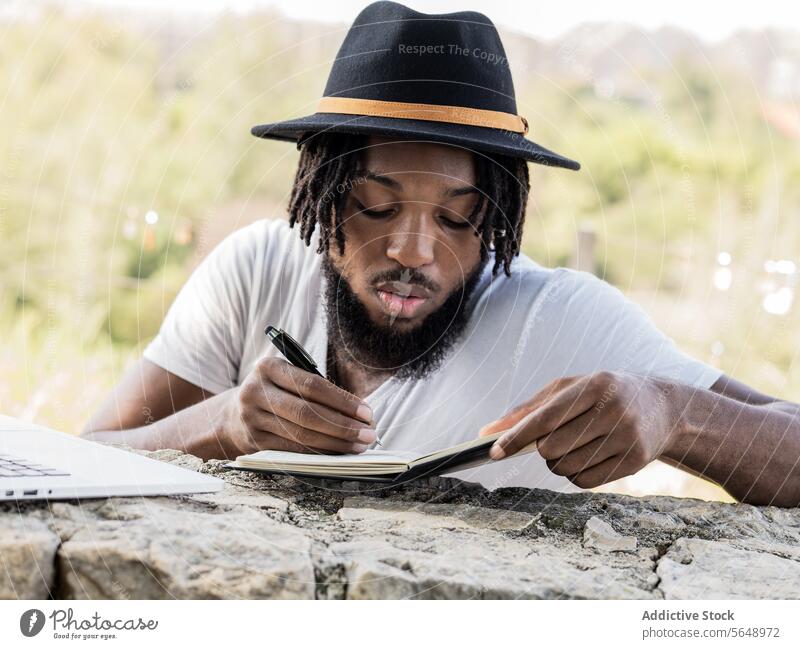 Focused black male podcaster writing notes on notebook man blogger park summer work write take note concentrate young african american ethnic dreadlocks beard