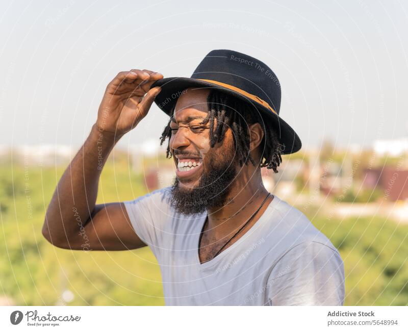 Cheerful black man adjusting hat in park fix sunlight laugh having fun joke humor funny touch smile positive cheerful male young african american ethnic