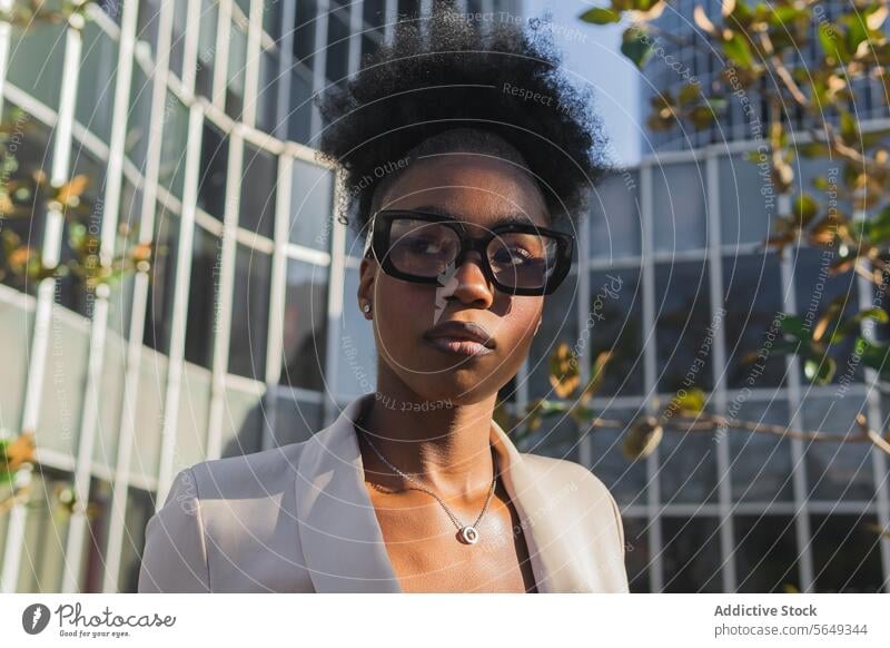 Serious black Businesswoman wearing glasses and blazer looking away while standing against building in city entrepreneur eyeglasses curly hair bun businesswoman