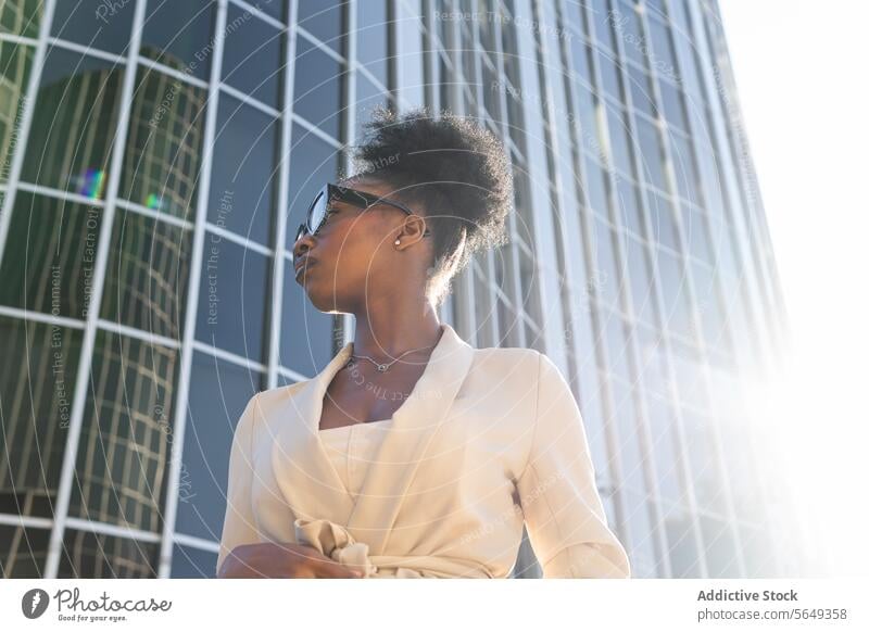 Low angle of black fashionable entrepreneur with eyeglasses looking away against building in city manager stylish low angle sunlight businesswoman curly