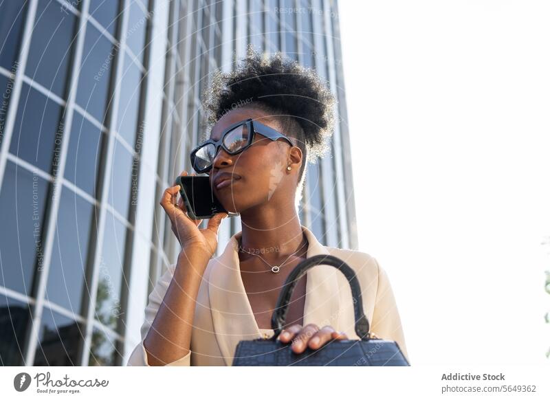 Low angle of Young black female manager talking on mobile phone sitting against building businesswoman smartphone purse urban city beautiful eyeglasses calm