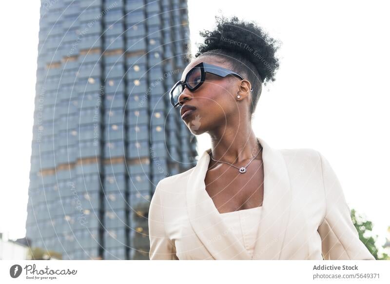 Low angle of Fashion black female entrepreneur in smart casual looking away while standing against building businesswoman fashionable eyeglasses bun low angle