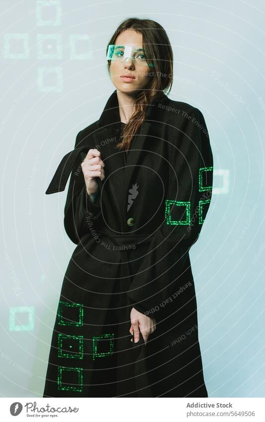 Portrait of young woman in smart futuristic VR glasses and long black trench coat standing looking at camera against illuminated background Woman Smart Glasses