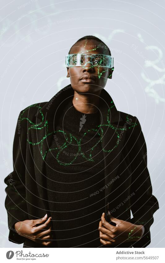 Portrait of confident young bold African American woman in smart futuristic VR glasses and long black trench coat standing looking at camera against neon light illuminated background