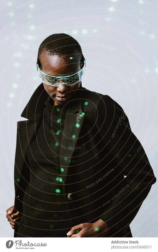 Portrait of confident young bold African American woman in smart futuristic VR glasses and long black trench coat standing with eyes closed against neon light illuminated background