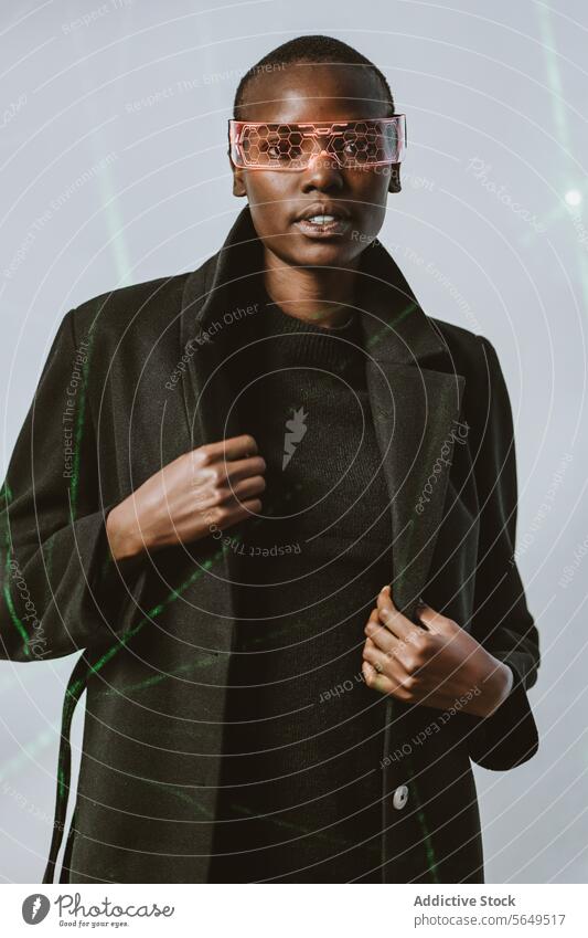 Portrait of confident young bold African American woman in smart futuristic VR glasses and long black trench coat standing looking at camera against neon light illuminated background