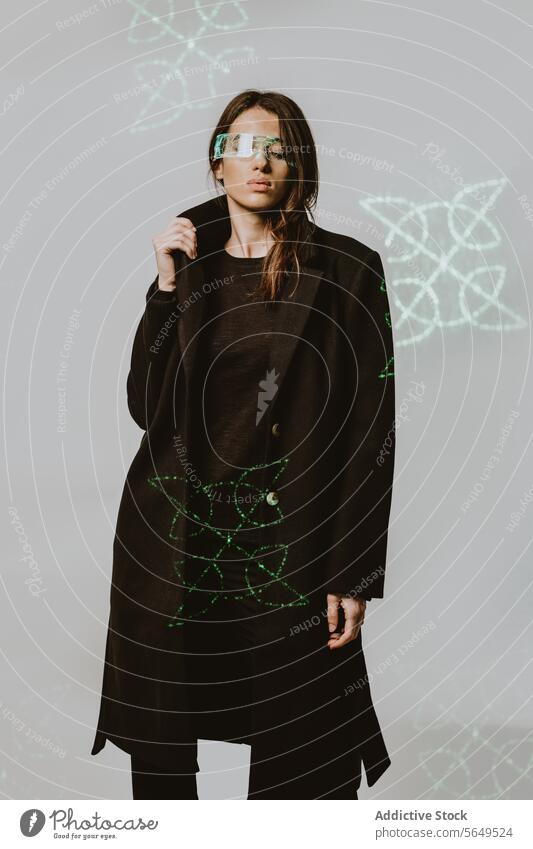 Portrait of young woman in smart futuristic VR glasses and long black trench coat standing with eyes closed against illuminated background Woman Smart Glasses