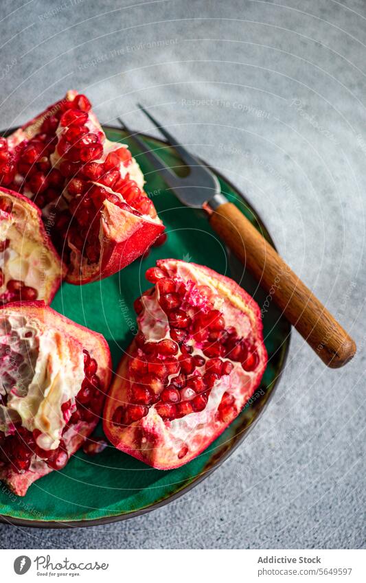 Fresh pomegranate pieces with vibrant red seeds on a green ceramic plate set against a textured grey concrete background fruit juicy ripe open section food