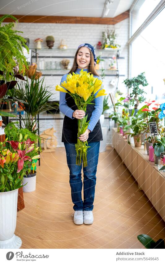 Smiling young female florist in casual clothes and bandana holding fresh yellow flowers while working in floristry during daytime woman owner arrange smile